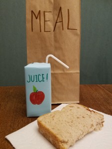 A brown paper bag labeled "meal" stands behind a juice box and a peanut butter sandwich on a napkin.