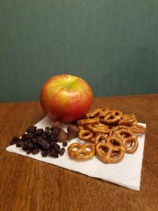 An apple, a pile of raisins, a pile of chocolate chips, and a pile of pretzels sit on a napkin.
